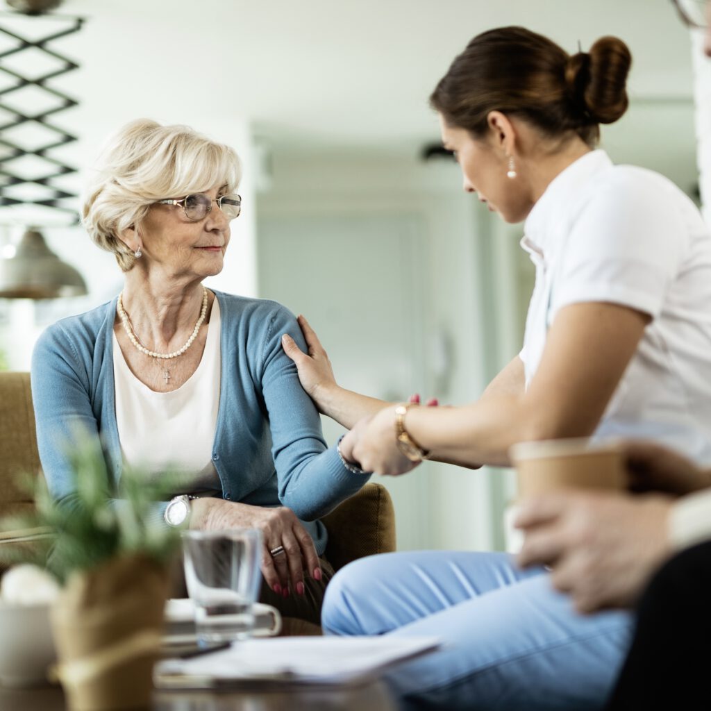 Female nurse communicating with senior woman while being in home visit. Focus is on senior woman.